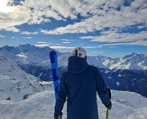 Skier standing on a snowy ridge in Verbier overlooking the Swiss Alps under a blue sky, holding skis and preparing to ski down the mountain.